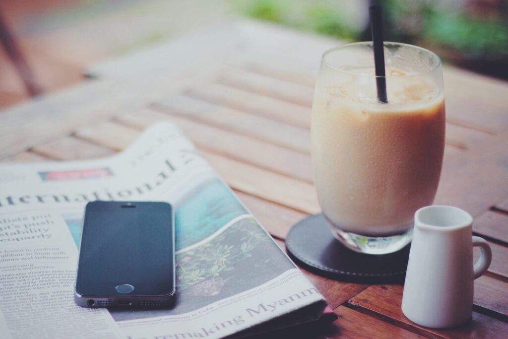 カフェオレと英字新聞の上に置かれたスマートフォンの写真